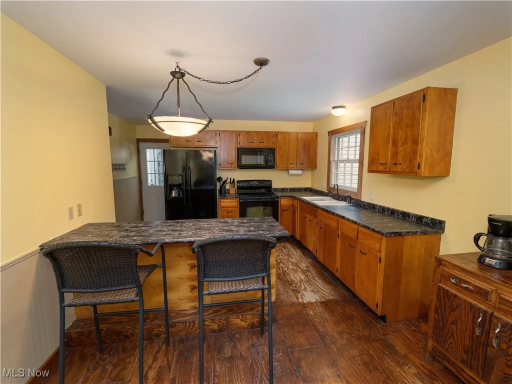 Kitchen featuring dark wood finished floors, black appliances, brown cabinetry, a kitchen bar, and hanging light fixtures