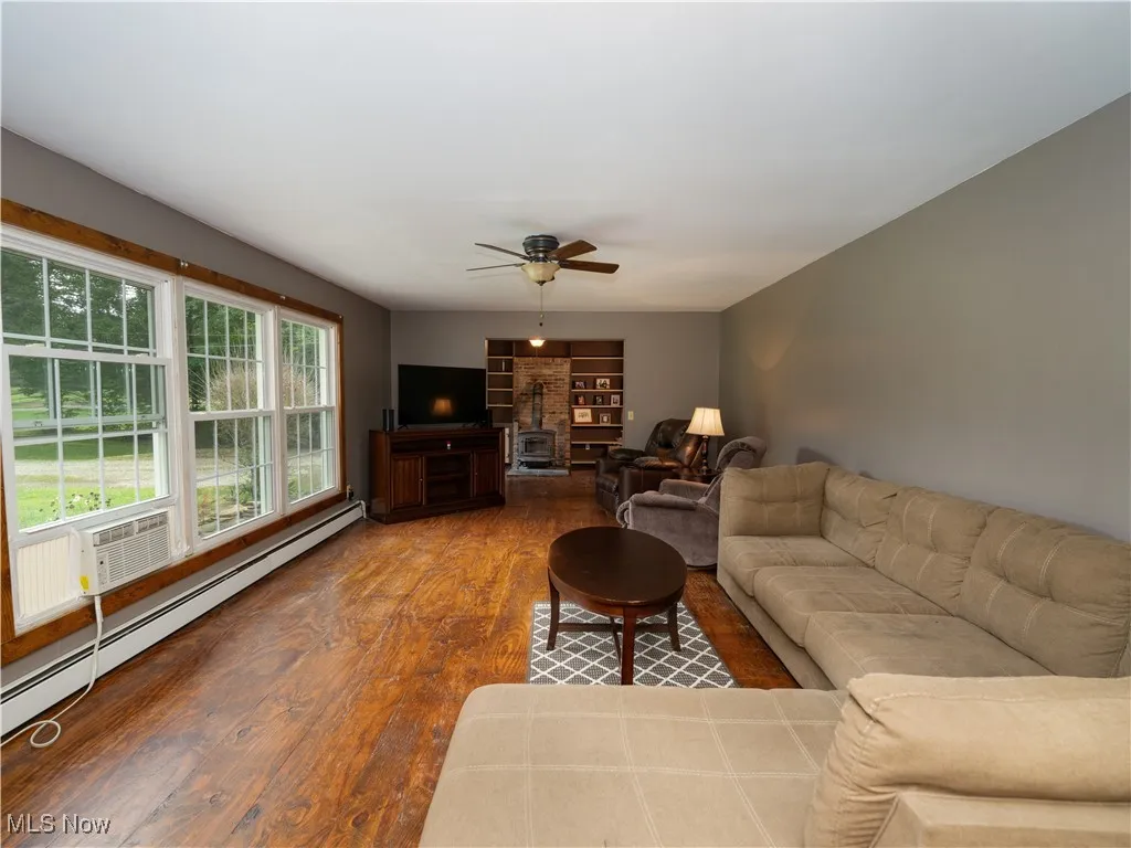 Living area featuring a wood stove, a baseboard heating unit, wood finished floors, and a ceiling fan