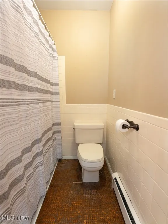 Bathroom featuring tile walls, a baseboard heating unit, a wainscoted wall, a shower with curtain, and dark tile patterned flooring