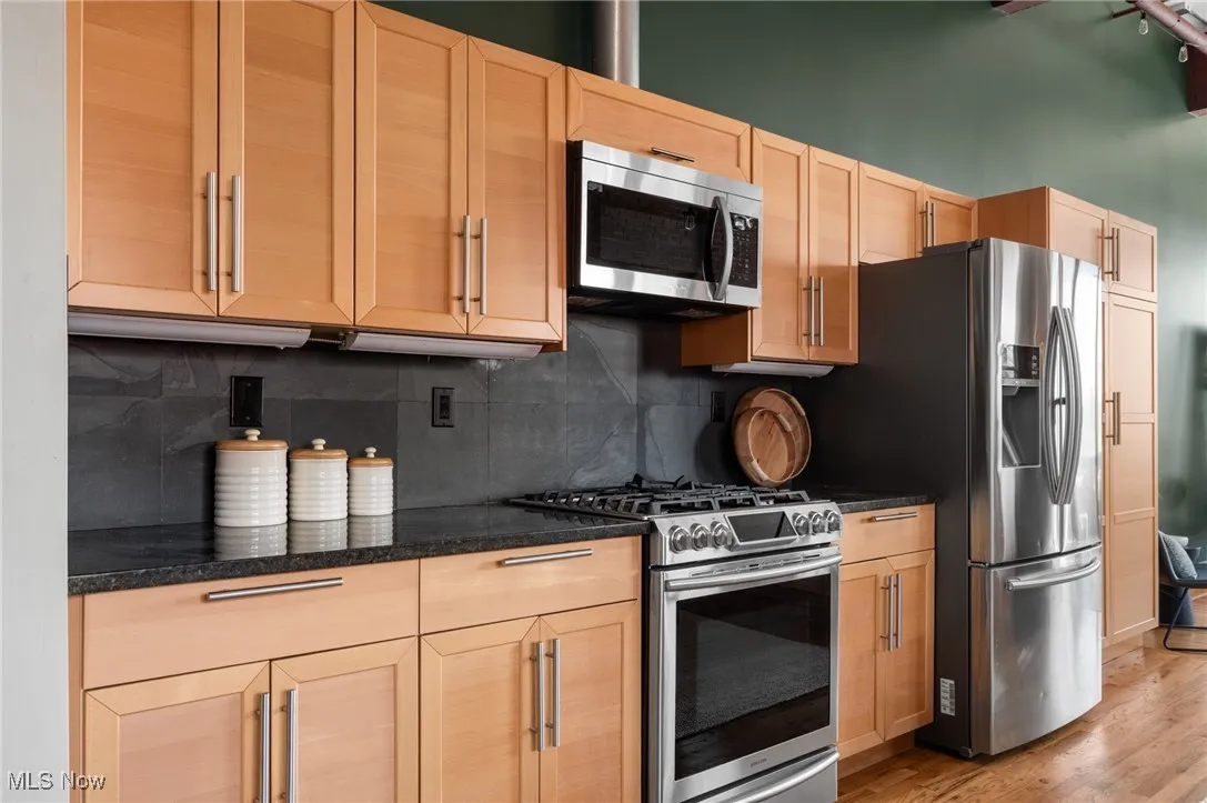 Kitchen with appliances with stainless steel finishes, backsplash, light brown cabinetry, light wood finished floors, and dark stone counters