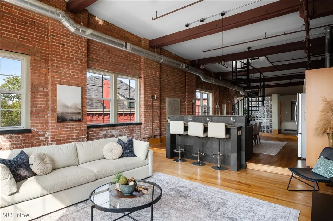 Living room with brick wall, light wood-style floors, beamed ceiling, and stairway