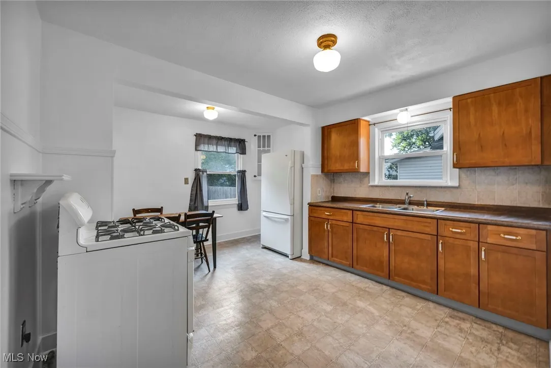 Kitchen featuring brown cabinetry, white appliances, dark countertops, backsplash, and light floors