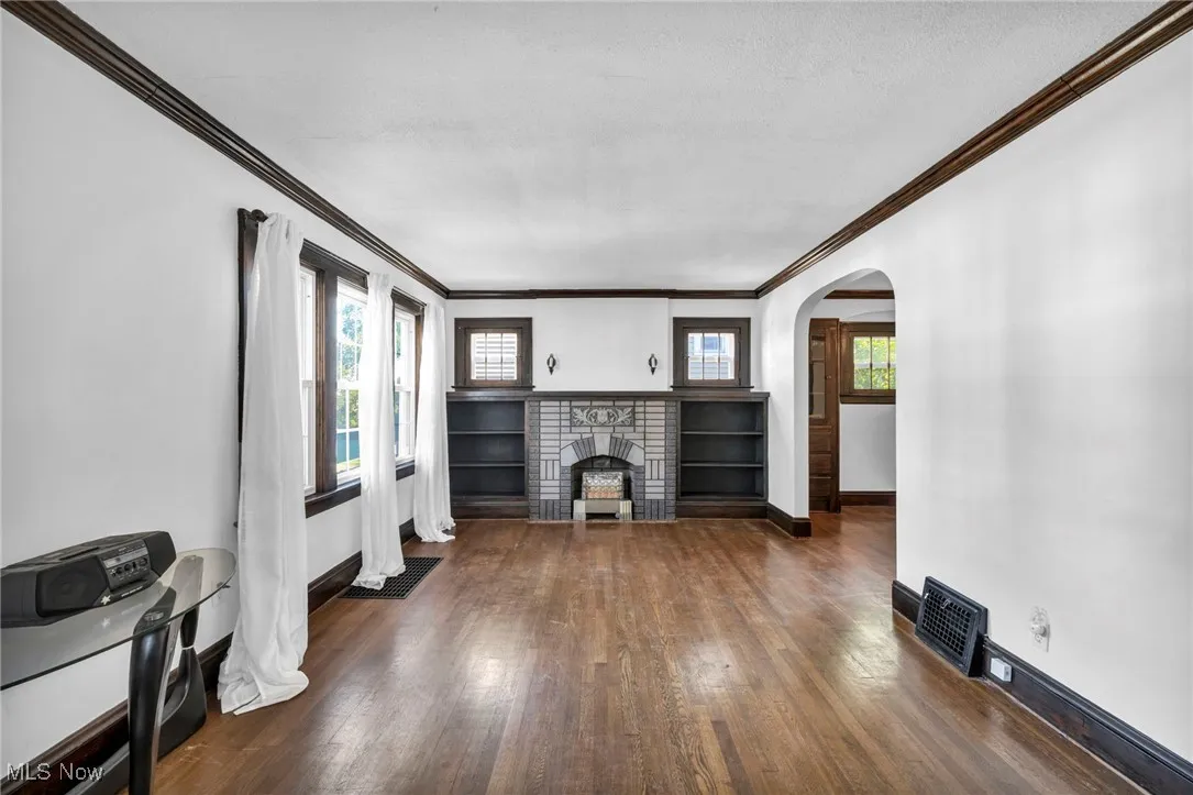 Unfurnished living room with healthy amount of natural light, dark wood-style floors, a brick fireplace, ornamental molding, and arched walkways