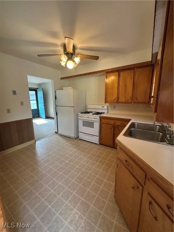 Kitchen with white appliances, light countertops, brown cabinetry, a wainscoted wall, and a ceiling fan