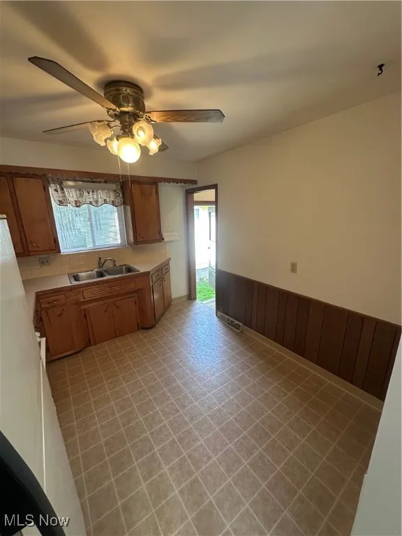 Kitchen featuring brown cabinetry, wainscoting, light countertops, a ceiling fan, and wood walls