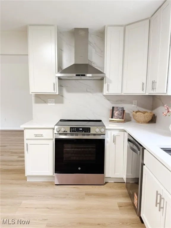 Kitchen featuring stainless steel appliances, wall chimney exhaust hood, white cabinets, light wood-style floors, and light stone counters