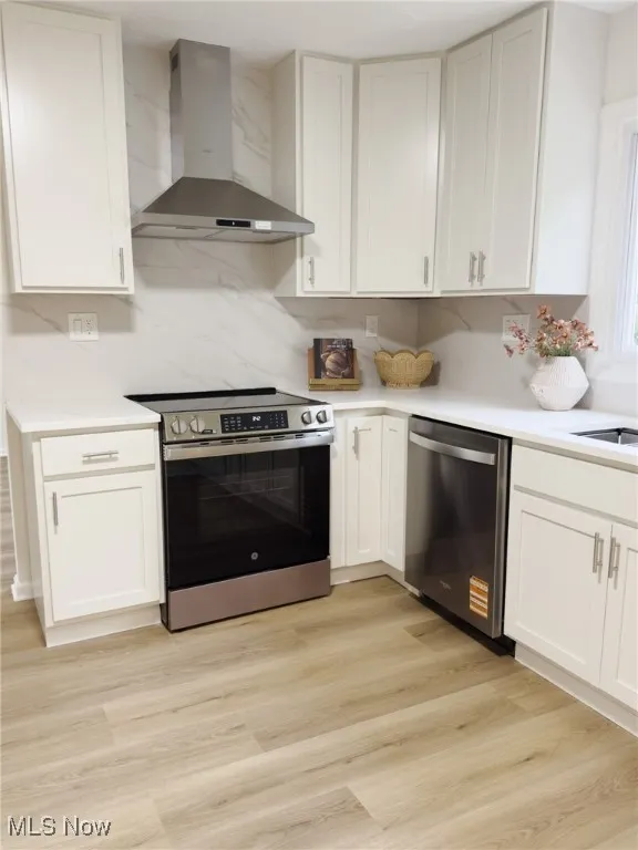 Kitchen with wall chimney exhaust hood, appliances with stainless steel finishes, light wood-style flooring, and white cabinetry