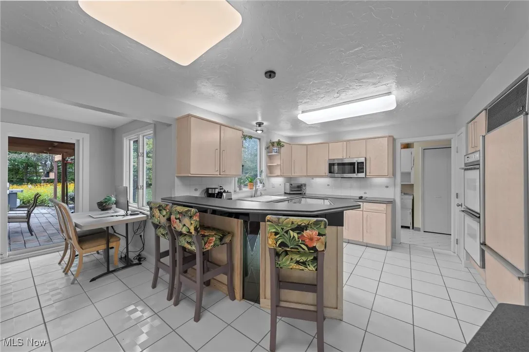 Kitchen featuring light brown cabinetry, a peninsula, light tile patterned floors, a textured ceiling, and dark countertops
