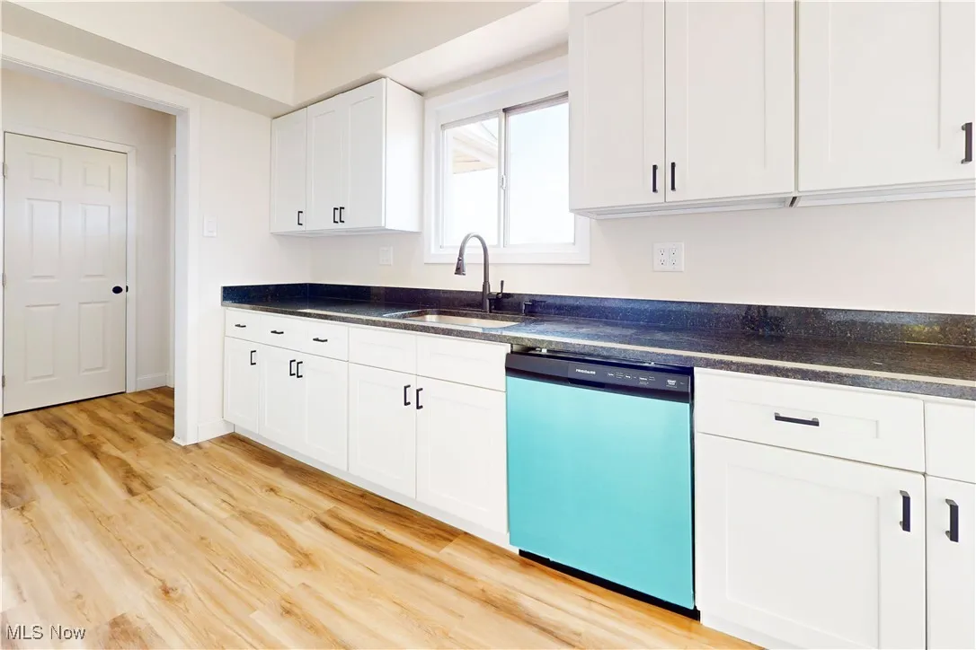 Kitchen with stainless steel dishwasher, white cabinets, light wood-type flooring, and dark stone countertops