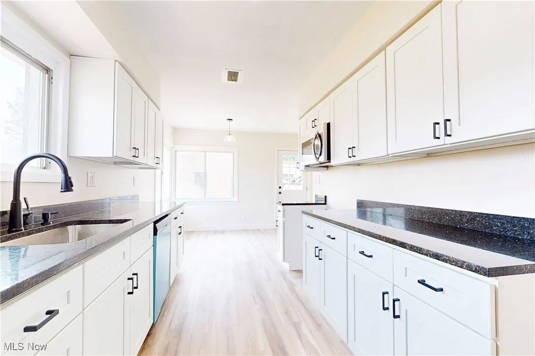 Kitchen with white cabinetry, light wood-style floors, appliances with stainless steel finishes, dark stone counters, and decorative light fixtures