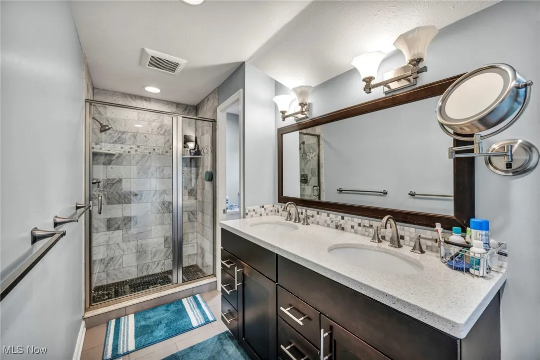 Full bath with light tile patterned floors, a shower stall, double vanity, and decorative backsplash