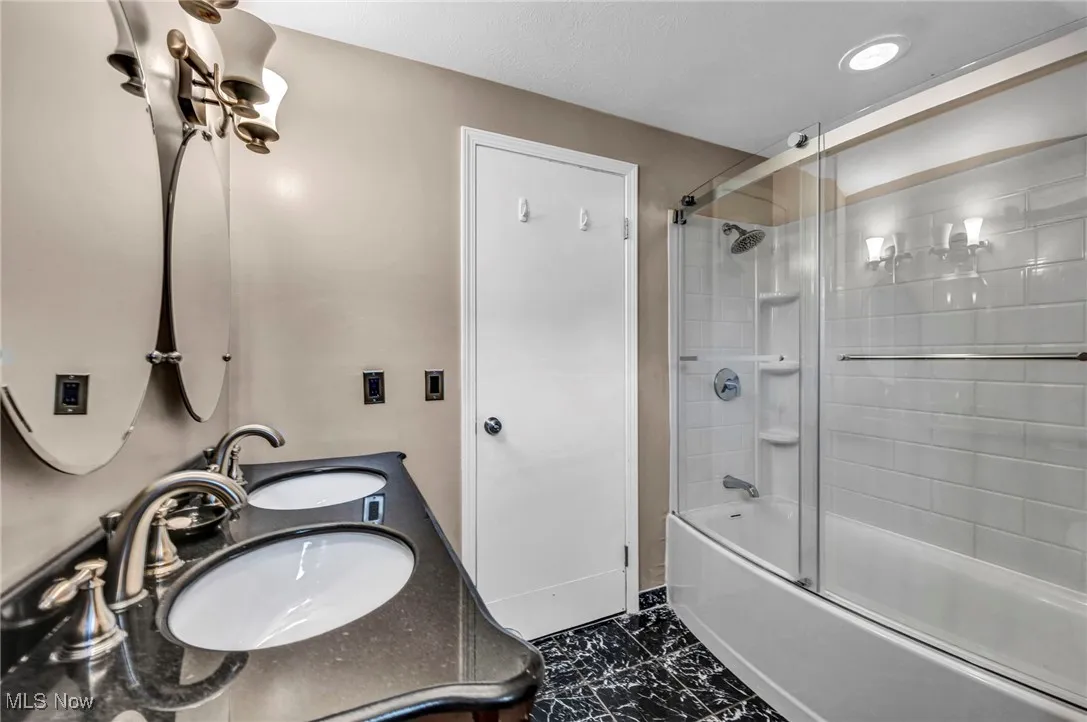 Bathroom with bath / shower combo with glass door, dark marble finish flooring, and double vanity