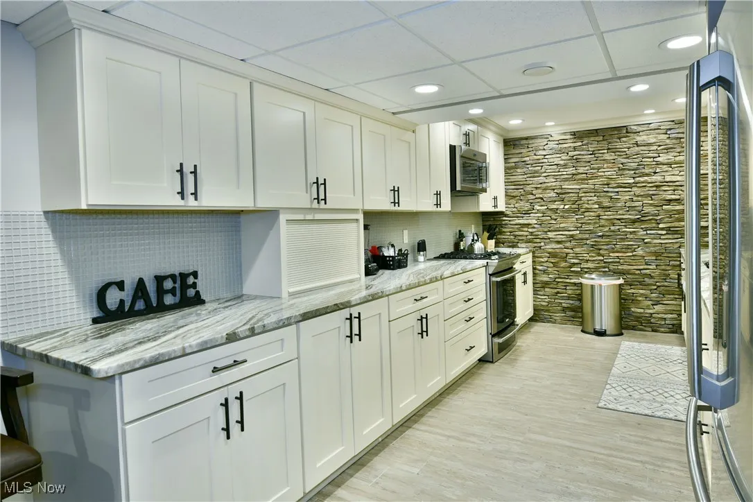 Kitchen featuring light stone counters, white cabinets, stainless steel appliances, a drop ceiling, and decorative backsplash