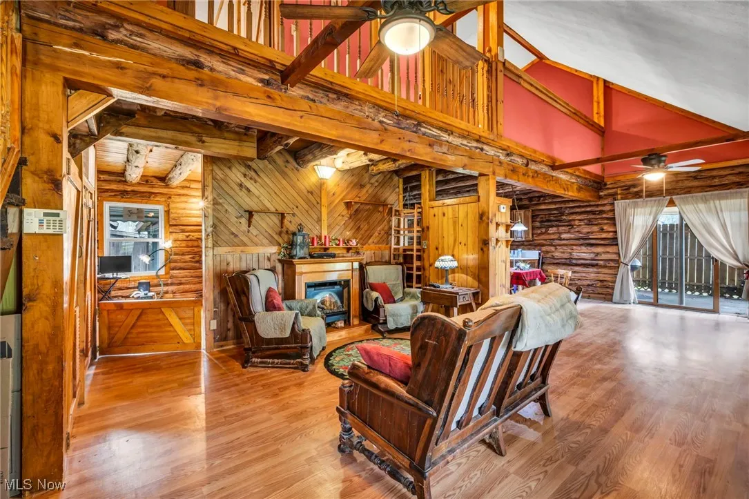 Living area featuring wood finished floors, a glass covered fireplace, wooden walls, ceiling fan, and beam ceiling