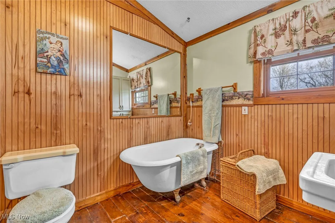 Bathroom featuring toilet, vaulted ceiling, a freestanding tub, wooden walls, and wood-type flooring
