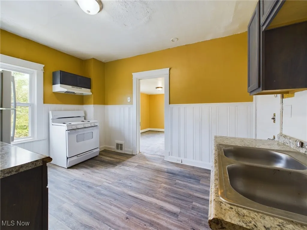 Kitchen featuring white appliances, wainscoting, dark wood-type flooring, under cabinet range hood, and dark brown cabinets