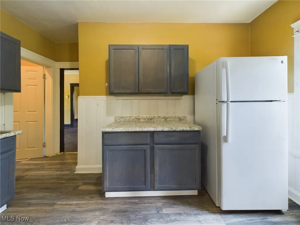 Kitchen with freestanding refrigerator, dark wood finished floors, wainscoting, dark brown cabinetry, and light stone countertops