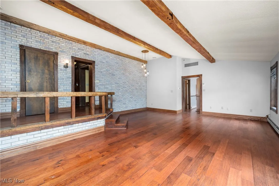 Unfurnished living room featuring wood finished floors, beam ceiling, brick wall, and a chandelier