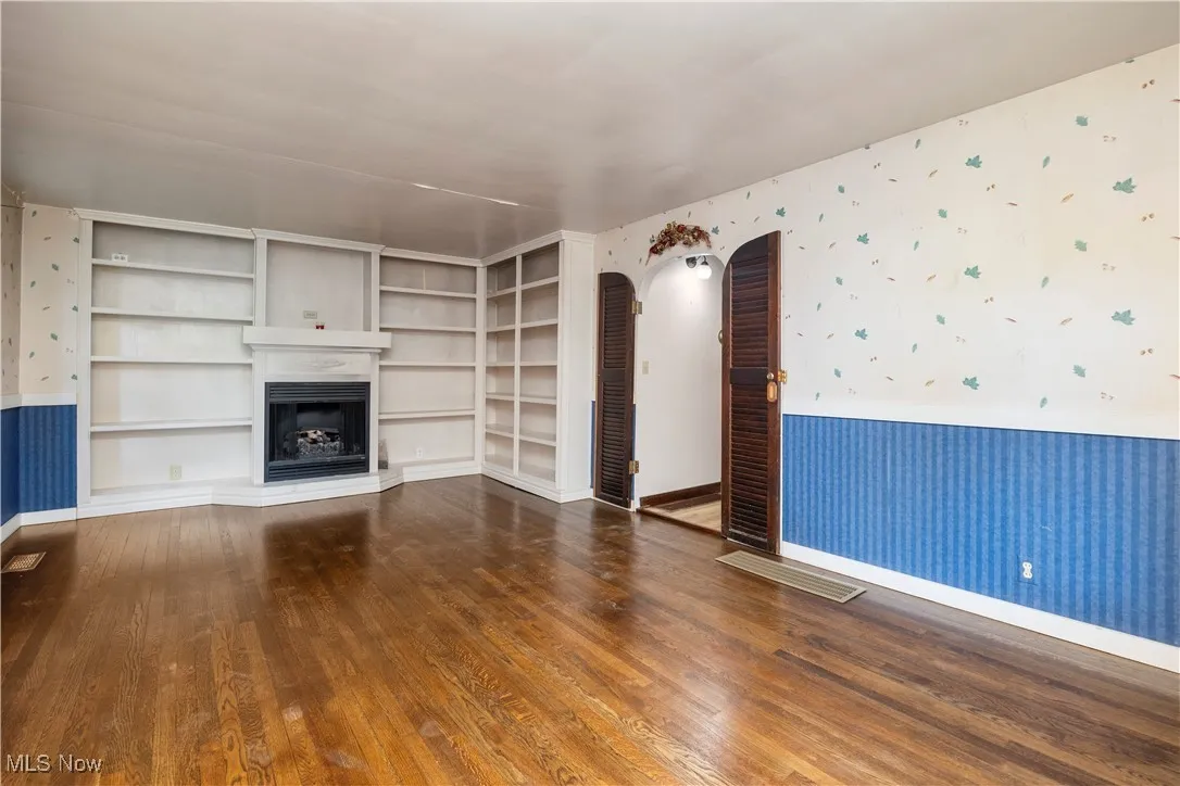 Unfurnished living room featuring wallpapered walls, wainscoting, built in features, arched walkways, and dark wood-style floors