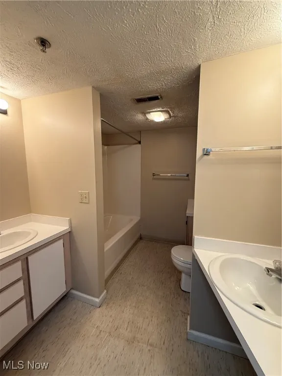 Full bath with a textured ceiling, two vanities, light flooring, and bathing tub / shower combination