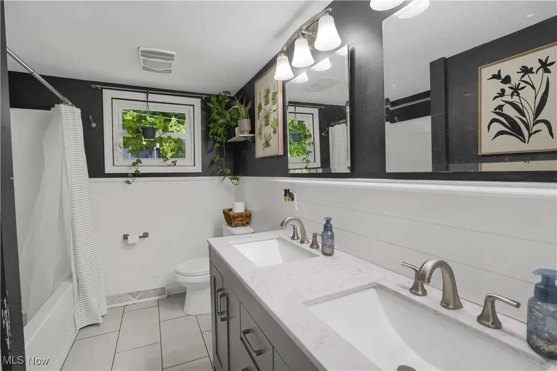 Full bath featuring a wainscoted wall, shower / bath combo with shower curtain, double vanity, light tile patterned floors, and tile walls