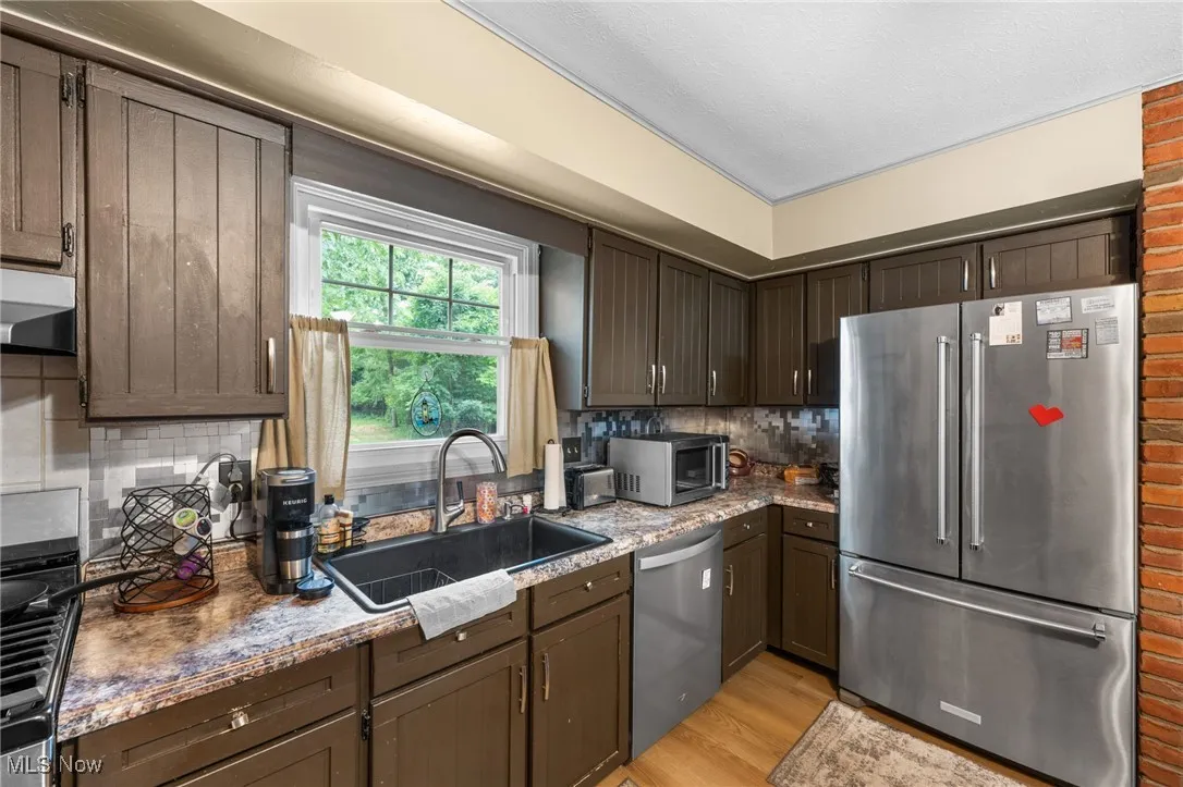 Kitchen featuring appliances with stainless steel finishes, dark brown cabinets, light wood-style floors, dark stone countertops, and tasteful backsplash