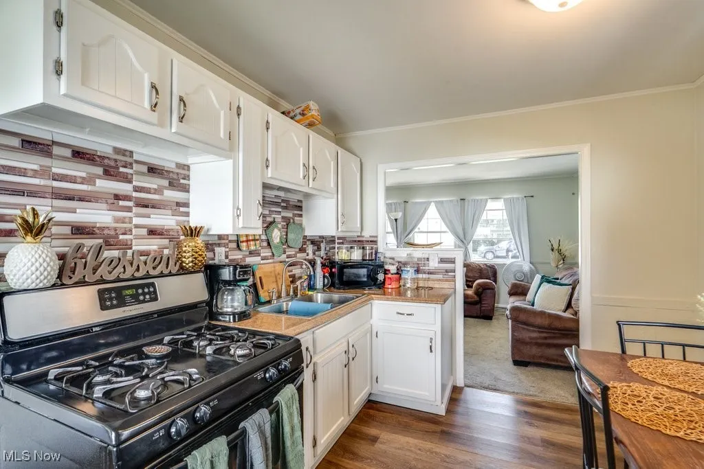 Kitchen featuring stainless steel gas range, decorative backsplash, dark wood-style flooring, ornamental molding, and white cabinetry