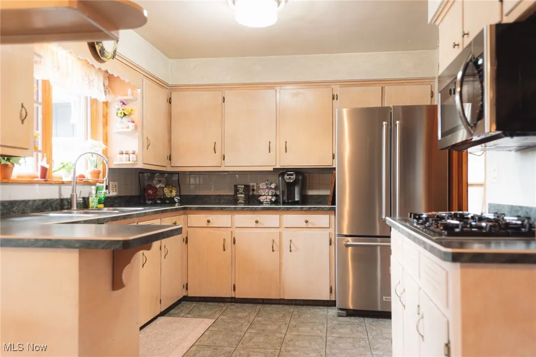 Kitchen with stainless steel appliances, dark countertops, light brown cabinetry, and decorative backsplash