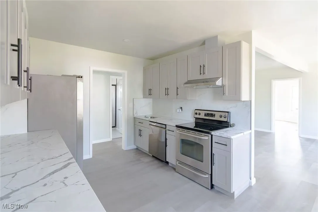 Kitchen with stainless steel appliances, light stone countertops, under cabinet range hood, backsplash, and white cabinetry