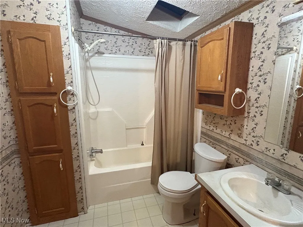 Full bath with wallpapered walls, a textured ceiling, shower / bathtub combination with curtain, light tile patterned floors, and vanity
