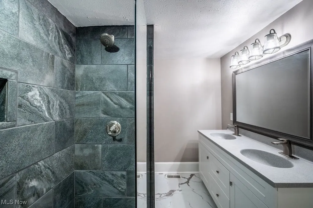 Full bathroom with a tile shower, a textured ceiling, light marble finish floors, and double vanity