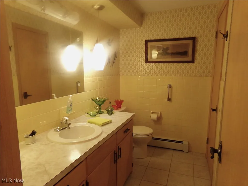 Bathroom featuring wallpapered walls, light tile patterned floors, vanity, baseboard heating, and tile walls