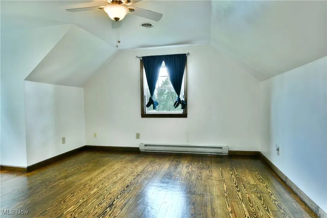 Bonus room featuring dark wood-type flooring, lofted ceiling, a baseboard radiator, and a ceiling fan