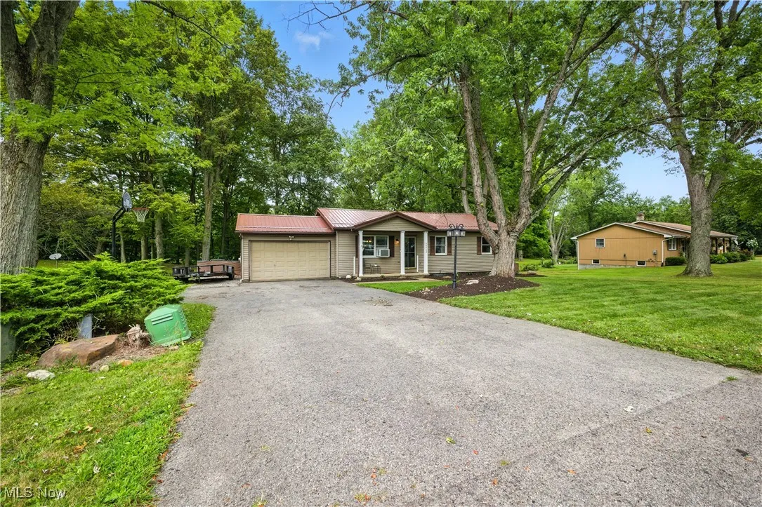 Single story home with an attached garage, driveway, a metal roof, a front lawn, and view of wooded area