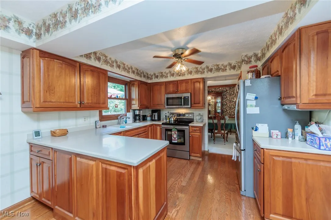 Kitchen with brown cabinetry, stainless steel appliances, light wood-type flooring, a peninsula, and wallpapered walls
