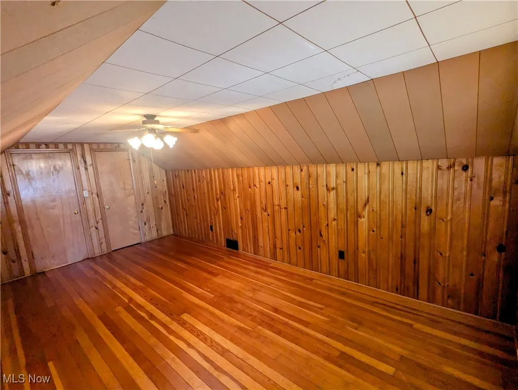 Another view of second floor bedroom with hardwood flooring, wooden walls, lofted ceiling, and ceiling fan