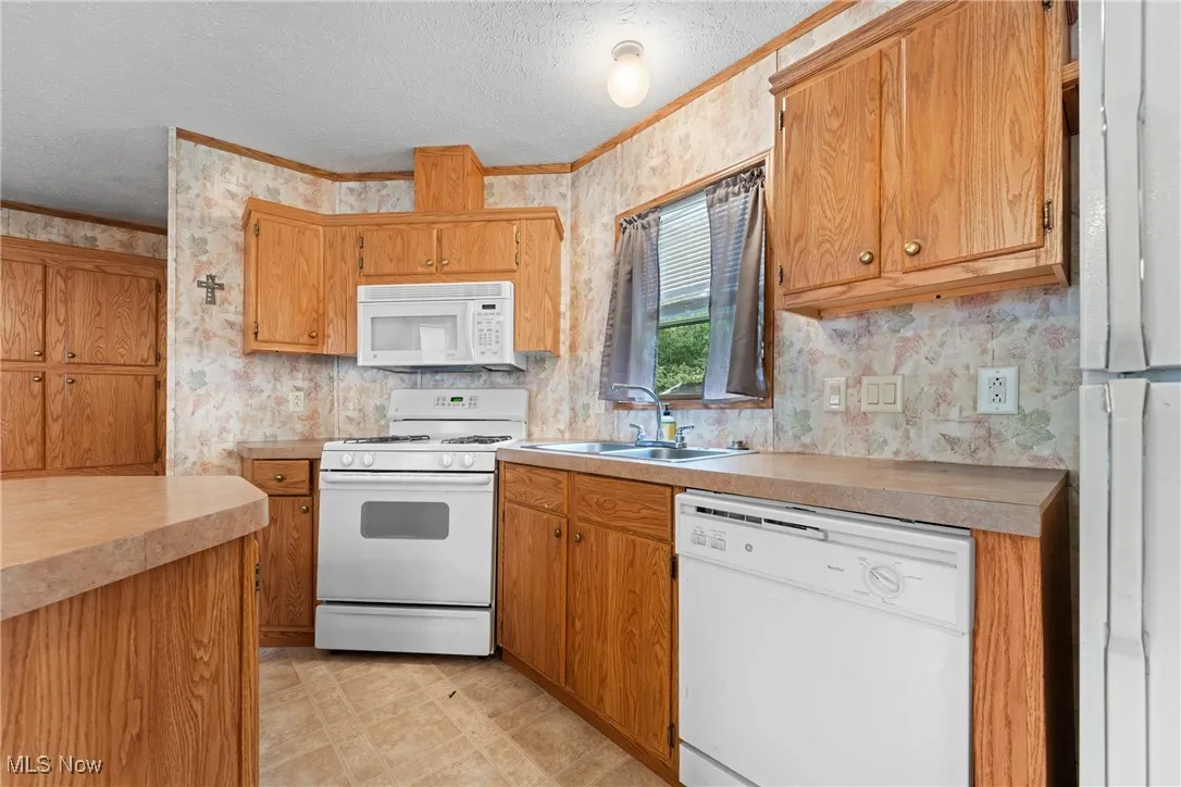 Kitchen featuring white appliances, wallpapered walls, light countertops, a textured ceiling, and brown cabinetry