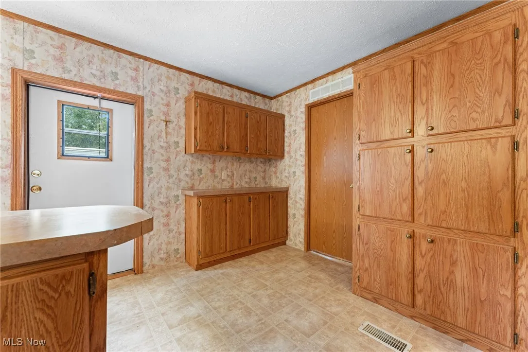 Kitchen with brown cabinets, ornamental molding, wallpapered walls, a textured ceiling, and light countertops
