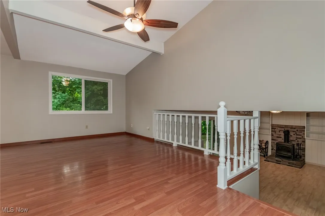 Unfurnished room with a wood stove, beam ceiling, light wood-type flooring, high vaulted ceiling, and ceiling fan