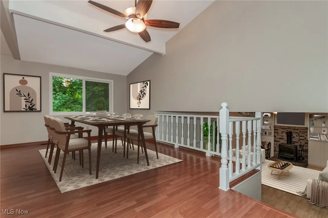 Dining area with a wood stove, wood finished floors, beam ceiling, a ceiling fan, and high vaulted ceiling