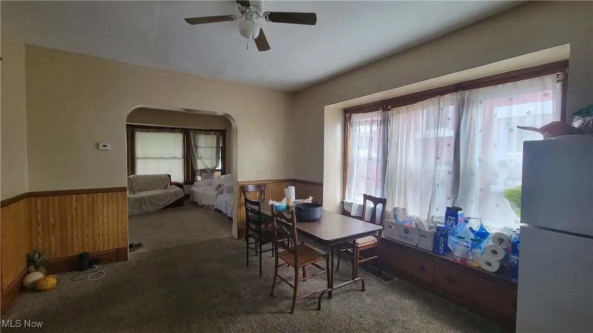 Dining space featuring wooden walls, wainscoting, dark carpet, arched walkways, and ceiling fan