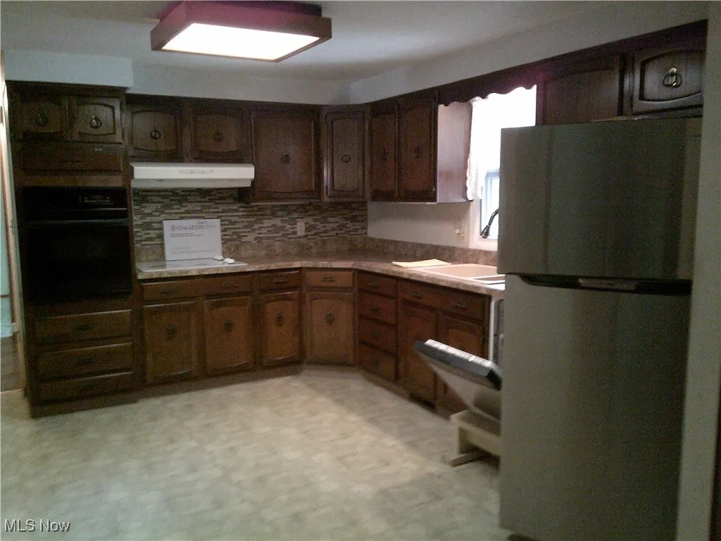 Kitchen with freestanding refrigerator, black oven, light floors, backsplash, and under cabinet range hood