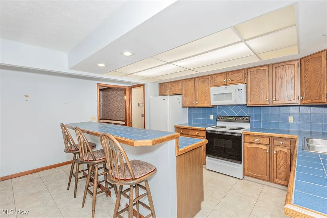Kitchen featuring brown cabinetry, a breakfast bar, white appliances, tasteful backsplash, and recessed lighting