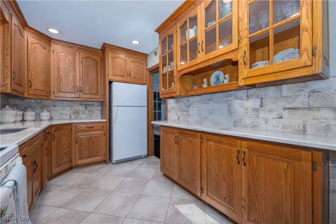 Kitchen with brown cabinetry, freestanding refrigerator, decorative backsplash, glass insert cabinets, and recessed lighting