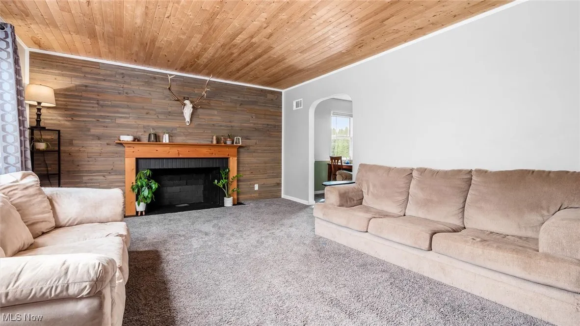 Carpeted living area featuring wood walls, a brick fireplace, arched walkways, ornamental molding, and wood ceiling