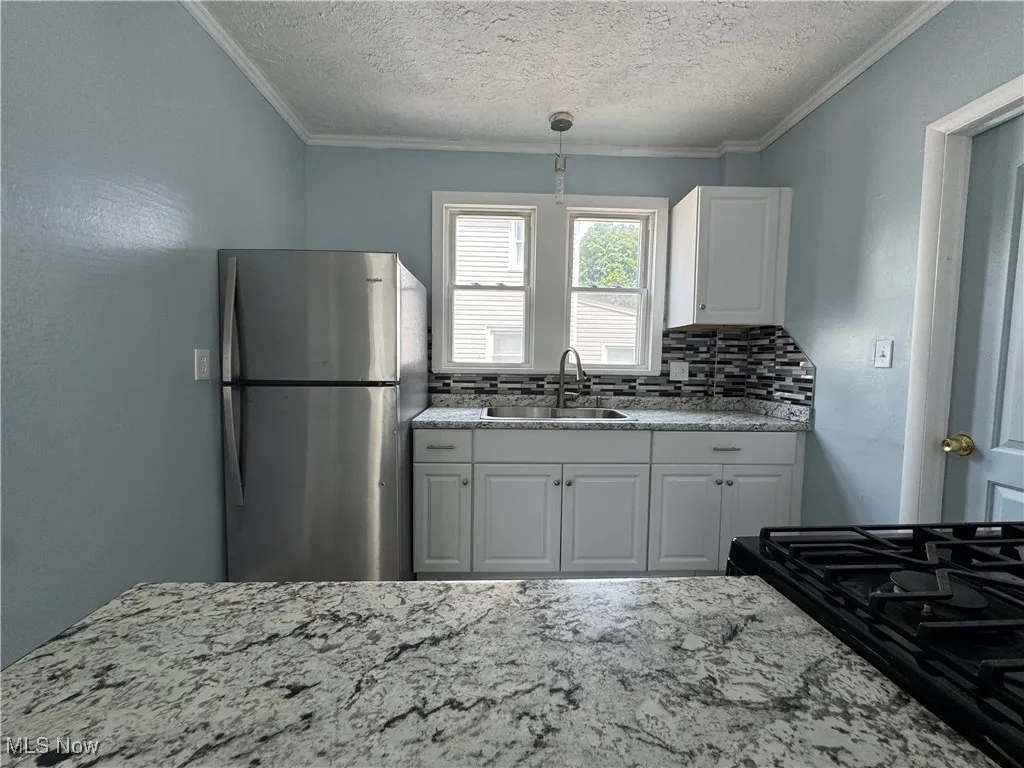 Kitchen with freestanding refrigerator, backsplash, light stone counters, decorative light fixtures, and ornamental molding