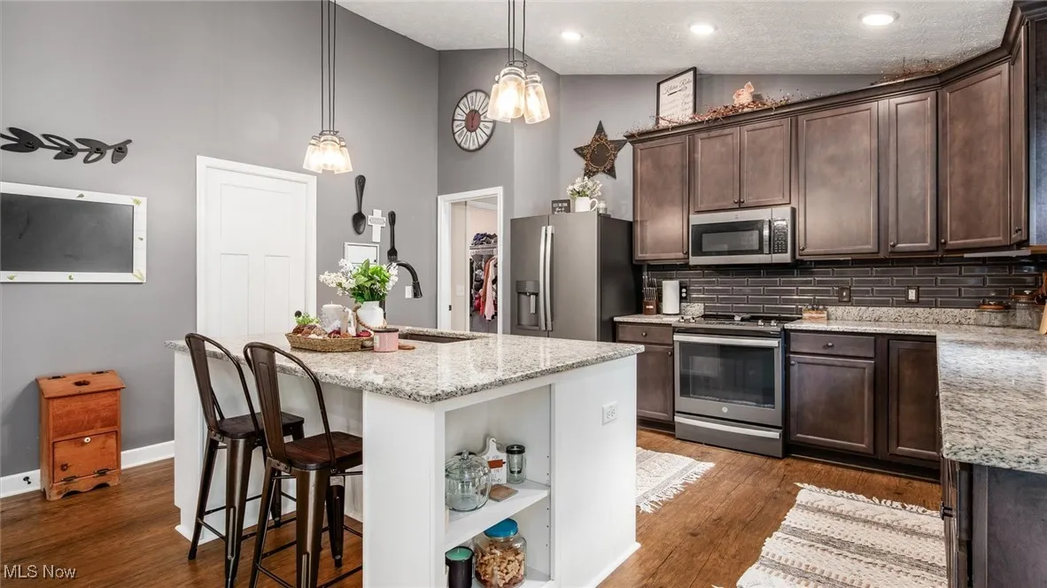 Kitchen with dark brown cabinets, stainless steel appliances, a kitchen island with sink, hanging light fixtures, and high vaulted ceiling