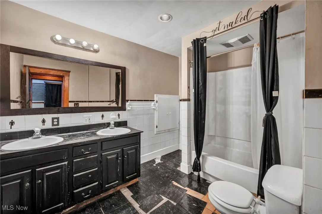 Full bath with shower / tub combo, double vanity, tile walls, and a wainscoted wall