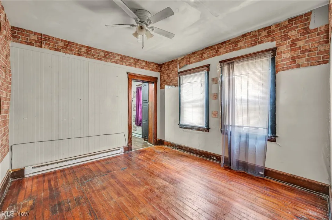 Unfurnished bedroom with wood-type flooring, brick wall, a baseboard heating unit, and a ceiling fan