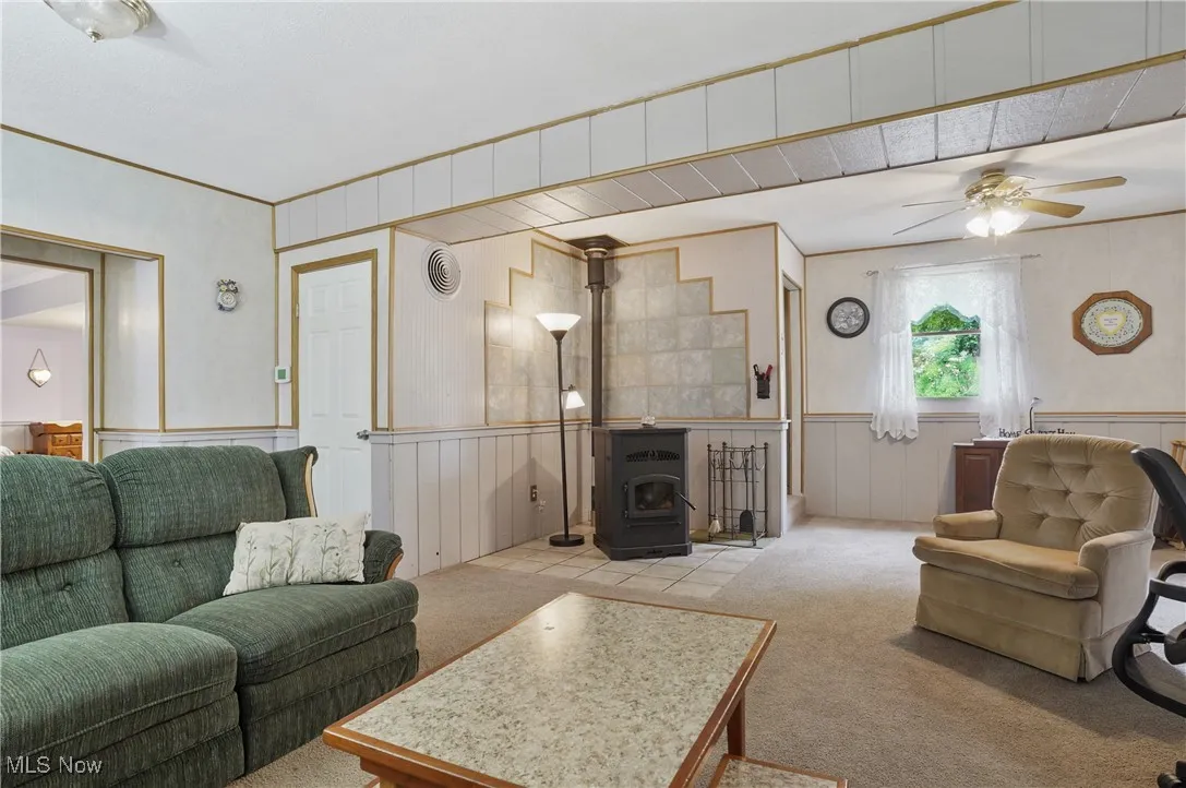 Living area with carpet floors, a wood stove, a ceiling fan, a wainscoted wall, and ornamental molding